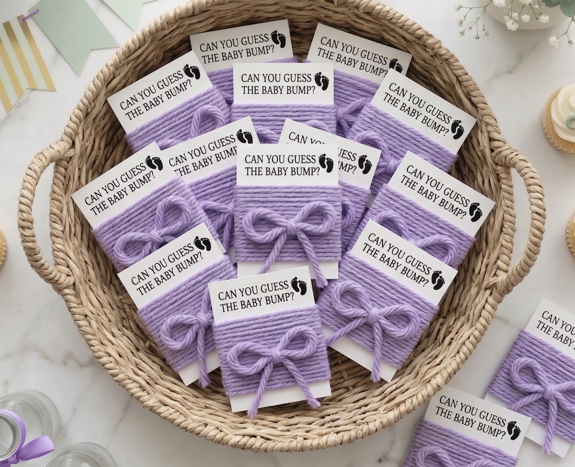 A woven basket filled with small lavender gift tags tied with purple ribbons. Each tag has a white label printed with the phrase 'CAN YOU GUESS THE BABY BUMP?' and a small black footprint icon.